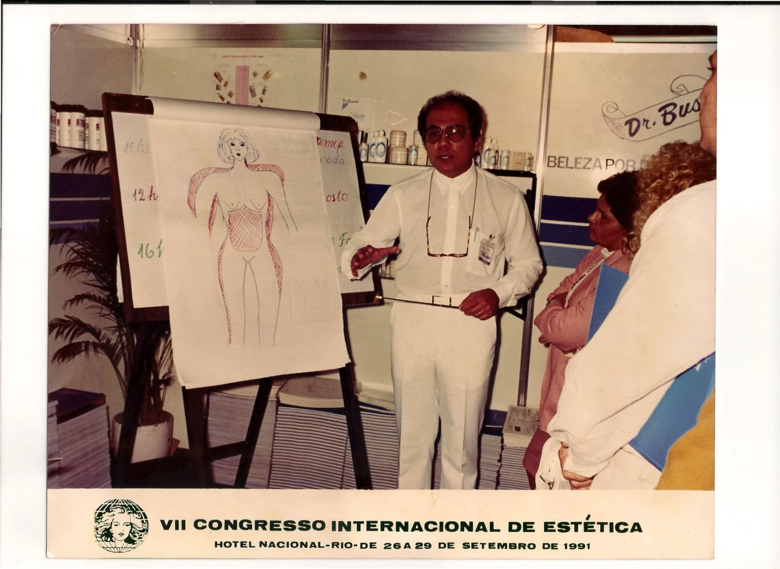 Dr. Bussade dando aula no stand, diante de um quadro com mapa corporal, em 1991